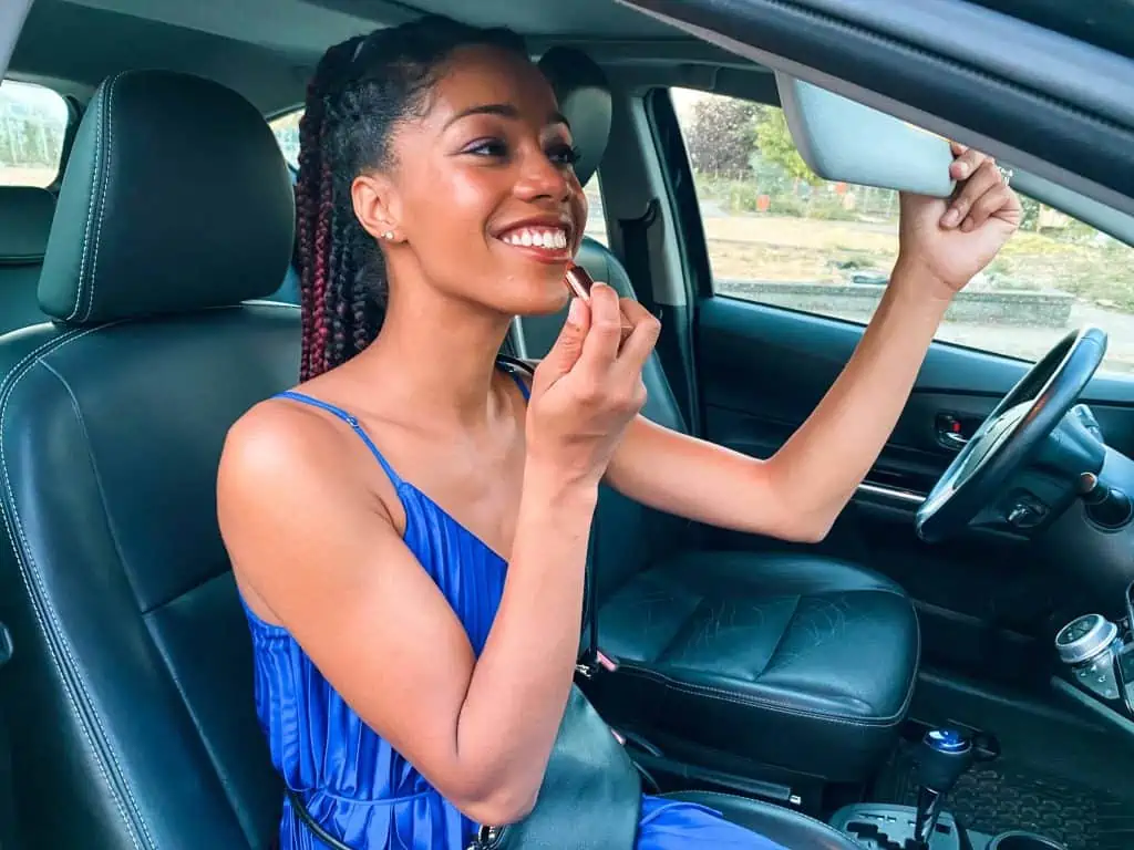 Black Blogger wearing Royal Blue Dress, applying lipstick while sitting in the passenger seat of an Evo Car Share in Vancouver