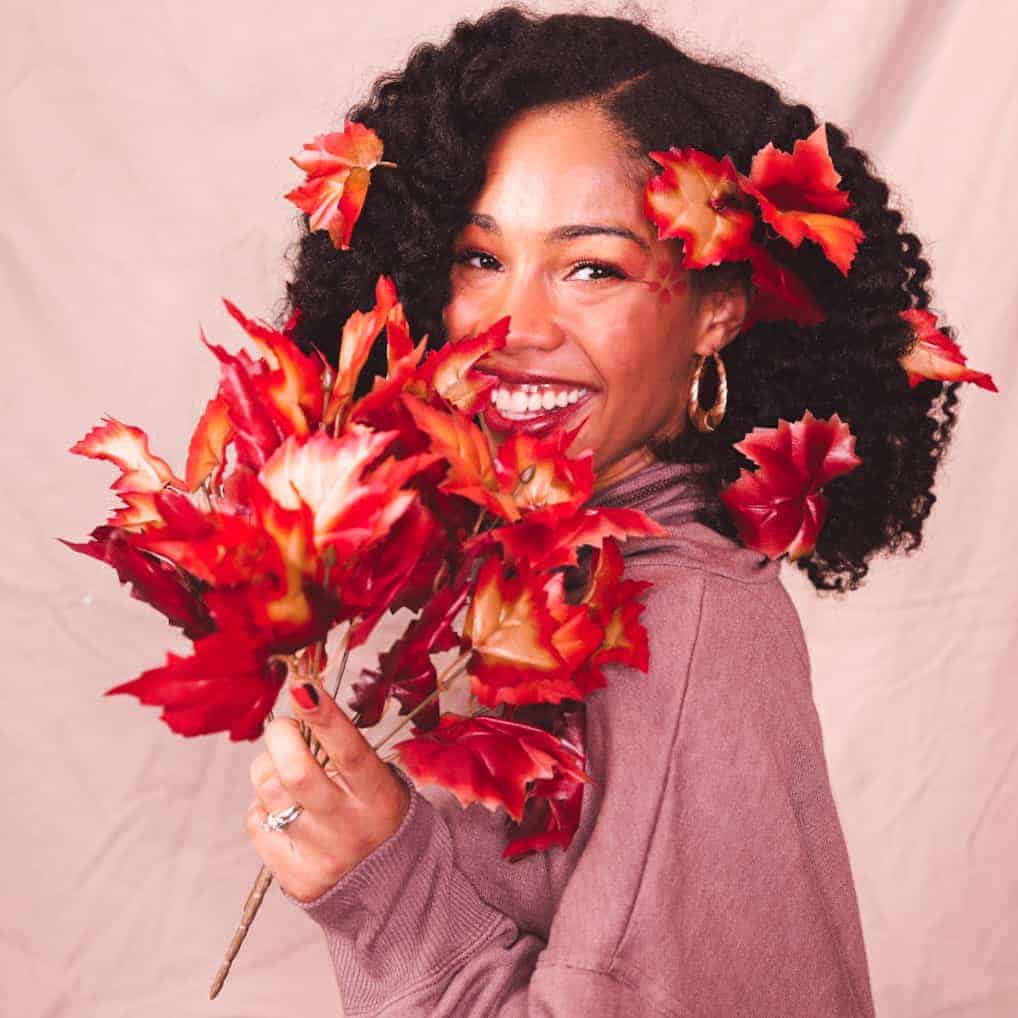 Black girl smiling covering face with rage and red fake fall leaves for DIY Halloween tree costume
