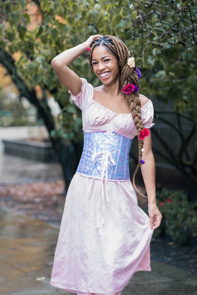 Black Woman ComfyGirlCurls smiling Dressed as Disney's Rapunzel for Halloween - Blonde Box Braids with Fake Flowers in hair, Pink Dres, Purple Corset